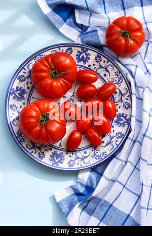 Verschiedene Arten reifer roter Tomaten und Kirschtomaten auf einem Teller mit Ziergegenständen. Saftige rote Tomaten in Blau und Weiß auf hellblauem Backen Stockfoto