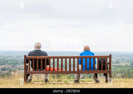 Blick von hinten auf zwei Männer in Wanderausrüstung und farbenfroher Kleidung, die auf einer Bank auf einem Hügel sitzen und einen Panoramablick über die Cotswolds-Landschaft haben Stockfoto