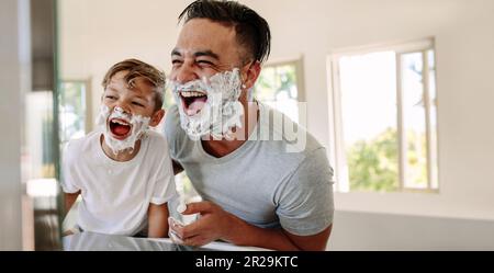 Vater und Sohn hatten Spaß im Bad, lachten fröhlich mit Rasierschaum im Gesicht. Junger alleinerziehender Vater, der sich einen Moment Zeit nimmt, um sich zu verbinden und Momente zu teilen Stockfoto