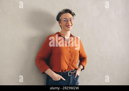 Porträt einer glücklichen Geschäftsfrau mit einer Brille, die vor der Kamera lächelt, während sie an einer grauen Wand steht. Reife Geschäftsfrau in Business-Casu Stockfoto
