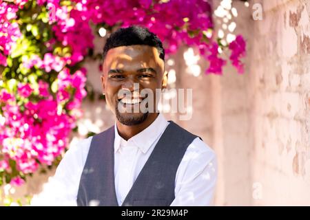 Lächelnder junger, afroamerikanischer Schwuler im Anzug, der gegen fröhliche Blüten und Mauern steht Stockfoto