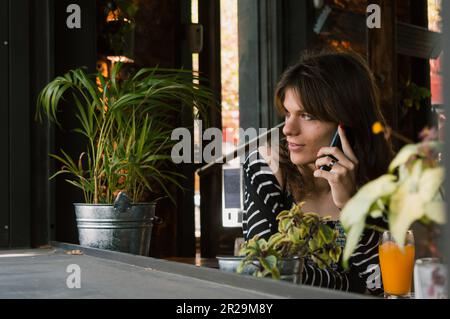 Junge lateinische Transgender-Frau argentinischer Herkunft, die telefoniert, im Restaurant sitzt, aus dem Fenster schaut und in den di schaut Stockfoto