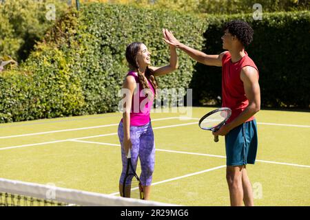 Glückliches, vielfältiges Paar mit Schlägern und High Fiving auf dem sonnigen Tennisplatz im Freien Stockfoto