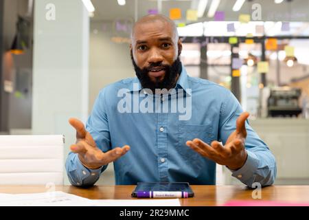 Porträt eines bärtigen afroamerikanischen Geschäftsmanns, der Gesten macht, während er im Büro am Schreibtisch sitzt Stockfoto