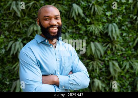 Porträt eines fröhlichen bärtigen afroamerikanischen Geschäftsmanns mit gekreuzten Armen, der gegen Pflanzen steht Stockfoto