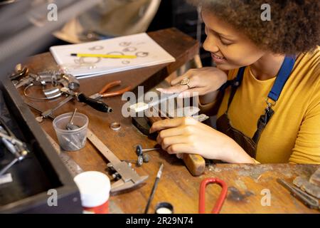 Glückliche birassische Mitarbeiterin, die Schmuck mit Werkzeugen in der Schmuckwerkstatt herstellt Stockfoto