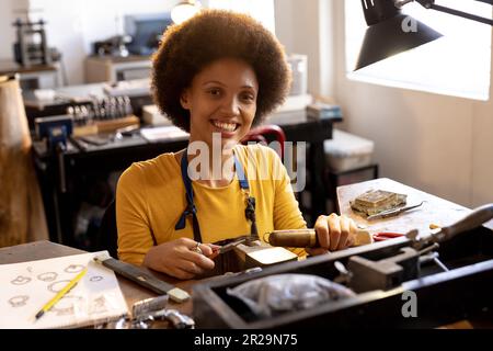 Porträt einer birassischen Arbeiterin, die Schmuck mit Werkzeugen herstellt und in der Schmuckwerkstatt lächelt Stockfoto