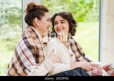 Entspannen Sie sich, lachen Sie, und lassen Sie sich von Mutter und Tochter mit Kaffeetasse zu Hause unterhalten, unterhalten Sie sich und knüpfen Sie sich zusammen. Glückliche Familie, Tee und auf der Veranda umarmt und rollt in einer warmen Decke Stockfoto