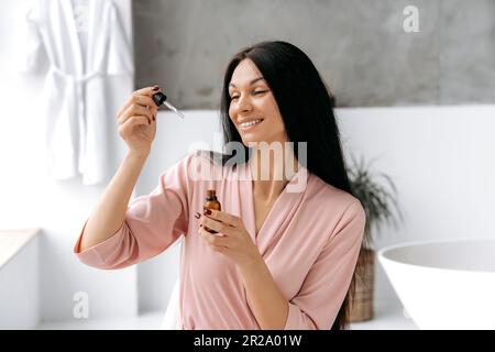 Schönheitskonzept. Charmante, gut gepflegte, weißbraune, langhaarige Frau sitzt im Badezimmer, hält in ihren Händen eine kleine Glasflasche mit Hyaluronsäure-Serum, verwendet Hautpflegeprodukte und lächelt Stockfoto