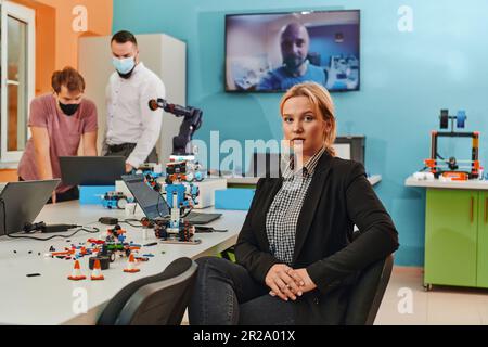 Eine Frau sitzt in einem Labor und löst Probleme und analysiert die Verifizierung des Roboters. Im Hintergrund unterhalten sich Kollegen online Stockfoto