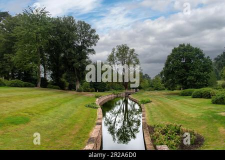 Genießen Sie die gepflegte Landschaft mit einem steinmauerten Fluss, der durch eine Wiese fließt, und Bäumen, die sich in der Ruhe des Wassers entlang des öffentlichen Fußwegs widerspiegeln Stockfoto