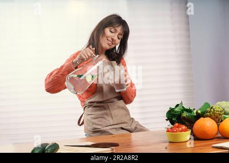 Eine weiße Frau in einer Küchenschürze sitzt an einem Küchentisch, wird von einem großen Fenster beleuchtet und gießt Gurkenwasser aus einem Krug in ein Glas. F Stockfoto