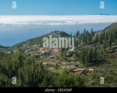 Blick aus der Vogelperspektive auf das kleine Dorf Igualero vom Gipfel des Alto de am Berg Garajonay. Weiße Wolken und die Insel El Hiero oben, blauer Himmel. La Gomera Stockfoto