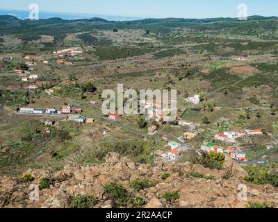 Blick aus der Vogelperspektive auf das kleine Dorf Chipude vom Gipfel des Tafelbergs Fortaleza. Weiße Wolken und die Insel La Palma oben, blauer Himmel. La Gomera, Kanarienvogel Stockfoto