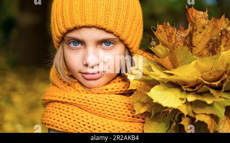 Wunderschönes kleines Mädchen in leuchtend gelbem, orangefarbenem Herbstkleid mit warmer Strickmütze und Schal, mit einem Strauß von Ahornblättern, Blättern im Wald. Lustiges Spiel Stockfoto