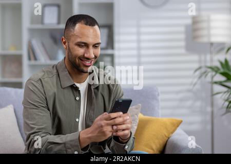 Ein junger afroamerikanischer Mann sitzt zu Hause auf der Couch und telefoniert. Schreibt eine Nachricht an ein Mädchen, chattet mit Freunden, liest Nachrichten, soziale Netzwerke. Nahaufnahme. Stockfoto