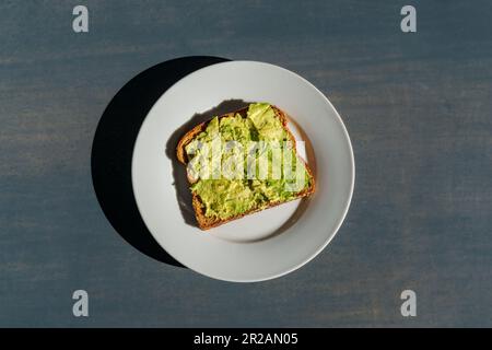 Avocado-Toast auf einem weißen Teller auf einer Betonarbeitsfläche in hellem Sonnenlicht mit harten Schatten Stockfoto