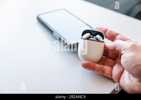 Kabellose Ohrstöpsel in der Ladetasche mit offenem Deckel, die von einem männlichen weißen Erwachsenen gehalten werden. Ein Smartphone befindet sich im Hintergrund auf dem Schreibtisch. Stockfoto