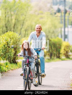 Porträts lächelnder Vater mit Tochter während des Sommerfahrens im Freien. Sie genießen die Zusammengehörigkeit im Sommer-Stadtpark. Glückliche Eltern und Chi Stockfoto