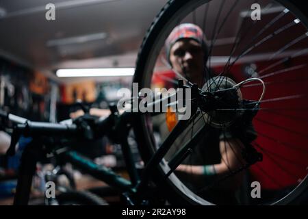 Detailaufnahme eines unbekannten, verschwommenen Mechanikers, der ein Motorrad mit einem Spezialwerkzeug repariert, der in einer Fahrradwerkstatt mit dunklem Innenraum arbeitet. Stockfoto