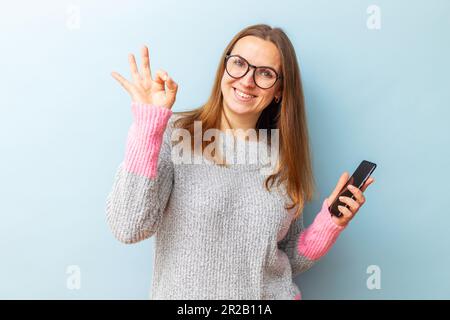 Glückliche brünette Frau mit Brille, die ein Smartphone in der Hand hält und eine gute Geste zeigt Stockfoto