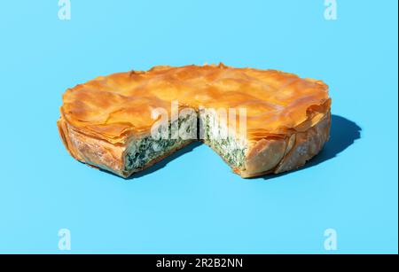 Hausgemachter balkan-Kuchen mit Fetakäse, Spinat und Phyllo-Teig, minimalistisch auf einem blauen Tisch. Geschnittener Spinatkuchen, isoliert auf blauem Hintergrund Stockfoto