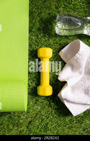 Fitness, Hydratation, Bewegung, gesunde und aktive Lebensweise Konzeptbild. Gerollte grüne Yogamatte, gelbe Kurzhantel und Wasserflasche auf grünem Gras i Stockfoto