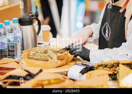 Die Küchenchefin kocht in der Schürze und Handschuhe schneiden frisch gebackenen Kuchen auf dem Markt. Straßenküche. , Selektiver Fokus Stockfoto