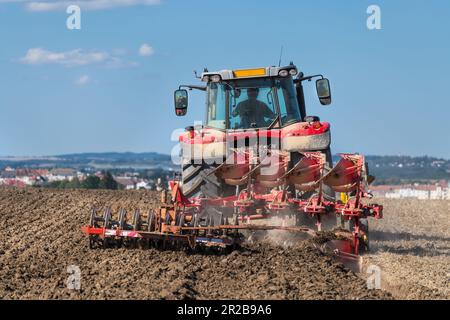 Roter Traktor auf dem Feld beim Pflügen und Eggen von Ackerland in der sommerlichen Natur unter blauem Himmel. Arbeitender Landwirt in landwirtschaftlichem Fahrzeug bei Bodenbearbeitung. Stockfoto