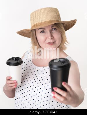 Blonde Frau, die schwarz-weißen Einwegbecher für Kaffee in verschiedenen Händen hält. Auswahlkonzept. Porträt der erwachsenen Frau mittleren Alters Stockfoto
