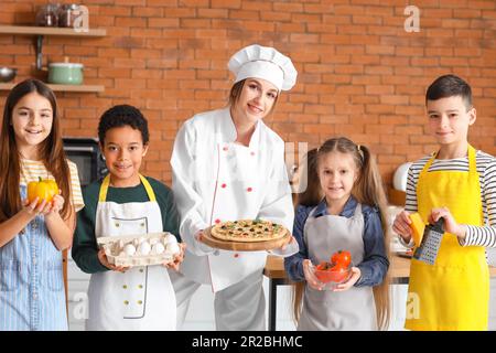 Weiblicher Koch mit zubereiteter Pizza und eine Gruppe kleiner Kinder nach dem Kochkurs in der Küche Stockfoto