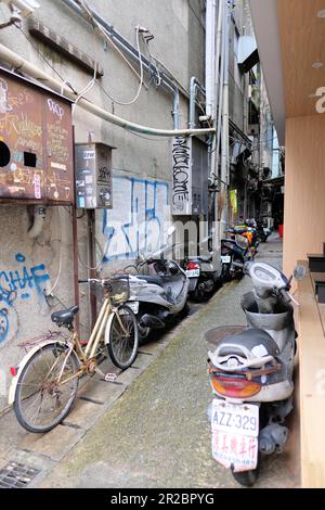 Enge Gasse zwischen den Gebäuden in Taipeh, Taiwan; geparkte Fahrräder und Motorroller; Graffiti und Aufkleber an der Wand und Sicherungskästen für elektrische Zähler. Stockfoto