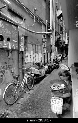 Enge Gasse zwischen den Gebäuden in Taipeh, Taiwan; geparkte Fahrräder und Motorroller; Graffiti und Aufkleber an der Wand und Sicherungskästen für elektrische Zähler. Stockfoto