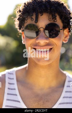 Porträt eines lächelnden, birassischen, fit sitzenden Mannes mit Weste und Sonnenbrille im sonnigen Garten Stockfoto