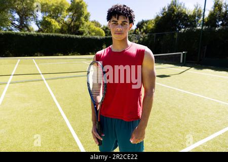 Porträt eines gut sitzenden, birassischen Mannes, der Tennisschläger auf einem sonnigen Tennisplatz hält Stockfoto