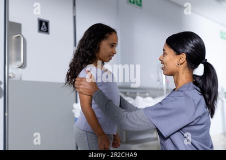 Glückliche birassische Ärztin und weibliche Patientin, die im Flur im Krankenhaus spricht Stockfoto