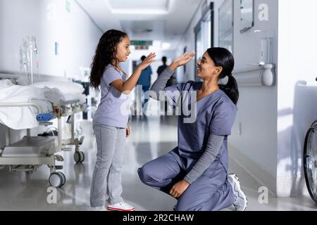 Happy knien birassische Ärztin und weibliche Patientin High Fiving im Flur des Krankenhauses Stockfoto