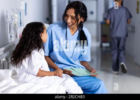 Glückliche birassische Ärztin und weibliche Patientin, die auf dem Bett sitzt und sich im Flur im Krankenhaus unterhält Stockfoto