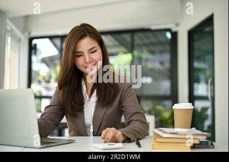 Wunderschöne, junge asiatische Buchhalterin, die mit dem Taschenrechner arbeitet und an ihrem Verkaufsbericht für Kunden an ihrem Schreibtisch im Büro arbeitet. Stockfoto