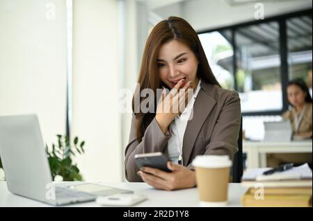 Aufgeregte und überraschte asiatische Geschäftsfrau, die eine unerwartete Nachricht auf ihrem Telefon erhält, während sie an ihrem Schreibtisch sitzt. Stockfoto