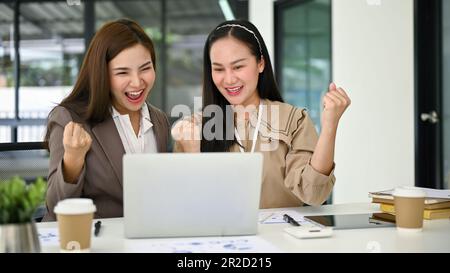 Zwei aufgeregte und fröhliche junge asiatische Geschäftsfrauen, die geballte Fäuste zeigen, sich freuen und ihren Projekterfolg gemeinsam im Büro feiern. Stockfoto