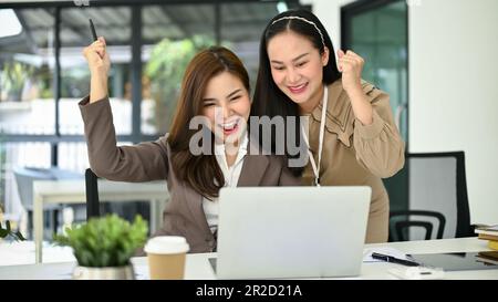 Zwei aufgeregte und fröhliche junge asiatische Geschäftsfrauen, die geballte Fäuste zeigen, auf einen Laptop-Bildschirm schauen, sich freuen und ihren Projekterfolg feiern Stockfoto