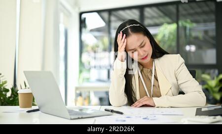 Gestresste und besorgte asiatische Millennials-Geschäftsfrau oder Buchhalterin, die während der Arbeit an Kopfschmerzen oder Migräne leidet. Stockfoto