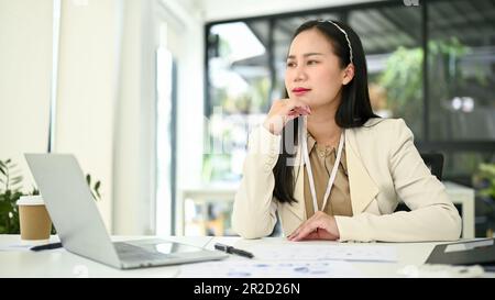 Rücksichtsvolle und gestresste asiatische Geschäftsfrau, die nachdenklich ist, aus dem Fenster blickt und beharrlich über einen Plan nachdenkt, um die Probleme zu lösen Stockfoto