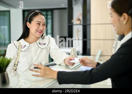 Eine lächelnde asiatische Kundin, die mit einer Empfangsdame spricht und ihre Unterkunft mit ihrer Kreditkarte in der Lobby bezahlt. Hospitality ser Stockfoto