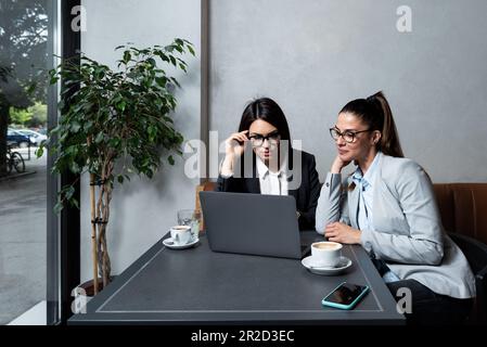 Zwei junge weibliche Geschäftspartner, Inhaber eines kleinen Unternehmens, neu auf dem Markt, eine Arbeitsunterbrechung in einem Café, reden und trinken Kaffee, machen pla Stockfoto