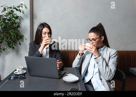 Zwei junge weibliche Geschäftspartner, Inhaber eines kleinen Unternehmens, neu auf dem Markt, eine Arbeitsunterbrechung in einem Café, reden und trinken Kaffee, machen pla Stockfoto