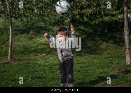 Glückliches Kind im Gras, das einen Fußball in die Luft wirft. Sportliches Kind in einem Park, das einen fliegenden Fußballball zwischenspeichert, während es in die Luft springt. Springer Junge Stockfoto