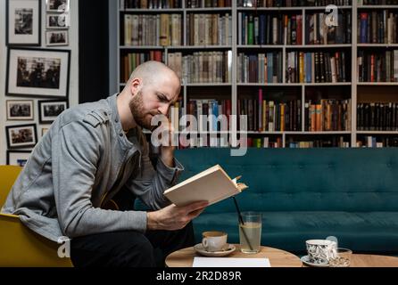 Kluger, seriöser, männlicher Kaukasier in Freizeitkleidung liest neuen interessanten Roman im Buchladen, erfolgreicher, fröhlicher Student, der ein positives Buch hält, während er sitzt Stockfoto