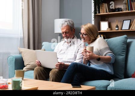 Glückliches Familienpaar mittleren Alters, das sich auf dem Sofa entspannt, mit dem Computer im Internet surft Informationen zu Hause einkauft. Älterer Mann, der Notebook-Apps zeigt Stockfoto